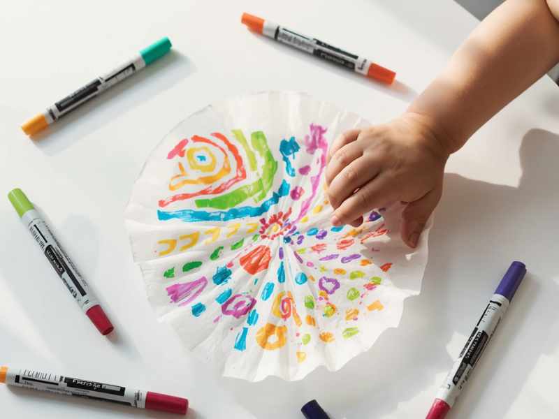 A bright, airy scene of a child’s hands coloring a flat coffee filter with vibrant washable markers on a white table, showing bold stripes, dots, and swirls in multiple colors, with extra markers scattered around.