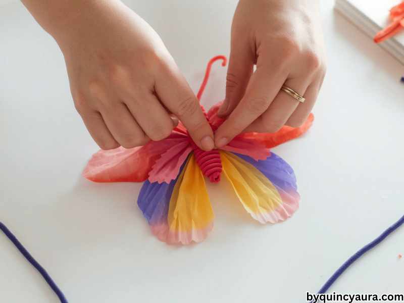 A bright, soft-lit close-up of hands shaping a colorful coffee filter into a butterfly by pinching the center and wrapping a pipe cleaner around it, with the wings fanned out beautifully in vibrant blended colors, set on a clean white table with extra pipe cleaners nearby.