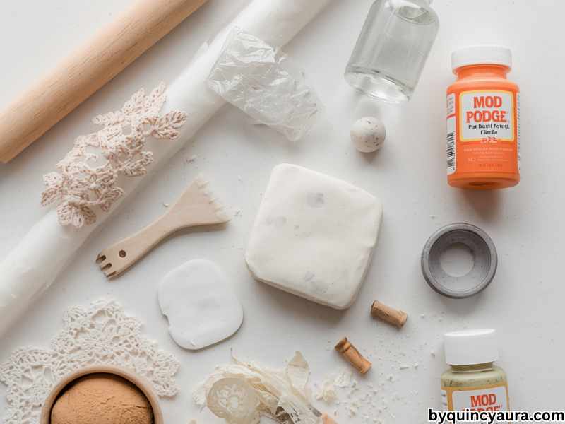 clean flat lay of air dry clay craft supplies arranged neatly on a white table, including white air dry clay, a rolling pin and glass bottle, a small bowl wrapped in plastic wrap, a plastic knife or clay tool, parchment paper, fine grit sandpaper, acrylic paints, paintbrushes, Mod Podge sealer, and optional textured items like lace, leaves, and stamps.